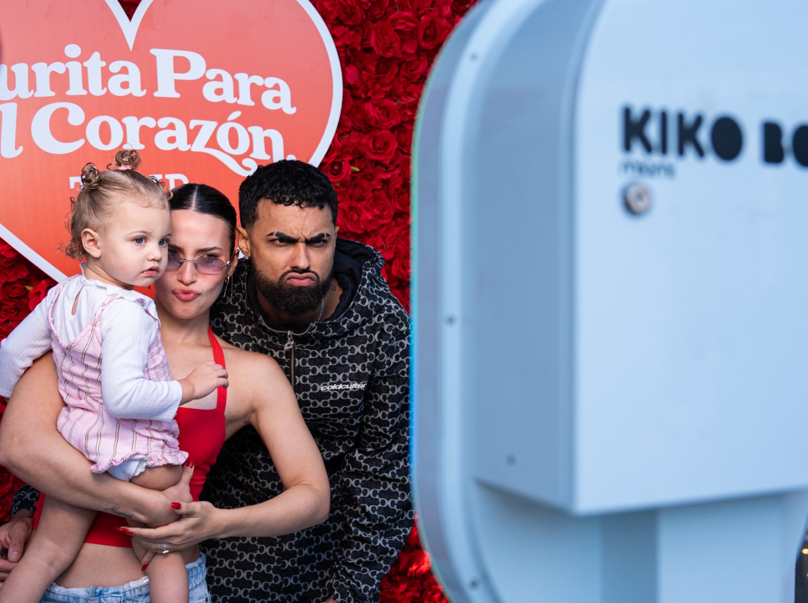Family posing at the ARO photobooth in front of a red rose wall at the Curita Para El Corazón tour